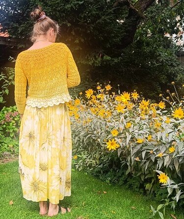 Person von hinten in gelbem Häkelpullover über hellgelbem, gemustertem Maxikleid vor blühenden gelben Blumen