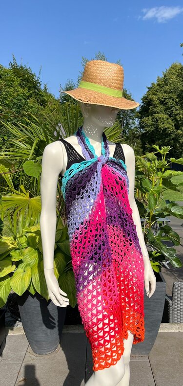 Mannequin trägt gehäkeltes Neckholder-Kleid in buntem Farbverlauf und Strohhut mit grünem Band, umgeben von Pflanzen und blauem Himmel.