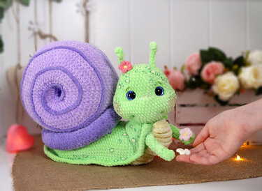 Plush crocheted amigurumi snail with textured green body, lavender spiral shell and blue eyes; a hand holds small white flowers near it.