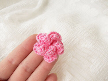 Hand hält kleine rosa gehäkelte Blüte mit fünf Blütenblättern vor hellem Stoff