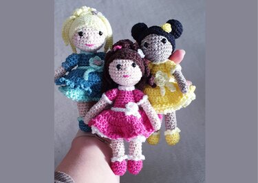 Hand hält drei kleine gehäkelte Amigurumi-Puppen in Rosa, Gelb und Blau mit Rüschenkleidern