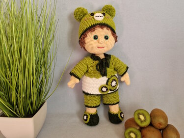 Gehäkelte Amigurumi-Puppe im grünen Kiwi-Outfit mit Bärenmütze; daneben liegen Kiwis und eine Topfpflanze.