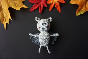 Gehäkelte graue Fledermaus‑Amigurumi mit großen Augen und ausgestreckten Flügeln, umgeben von gelben, roten und orangefarbenen Herbstblättern auf dunklem Hintergrund.