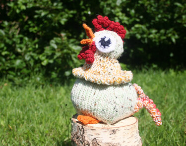 Hellgrünes gestricktes Amigurumi-Huhn mit rotem Kamm und orangefarbenem Schnabel auf Holzstumpf vor grünem Hintergrund.