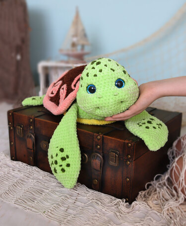 Green crochet sea turtle amigurumi with blue eyes on a wooden trunk, hand supporting its head
