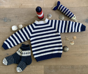 Blau-weiß gestreifter Kinderpullover und passende Anker-Socken auf hellem Holzboden, umgeben von Muscheln und maritimer Deko.
