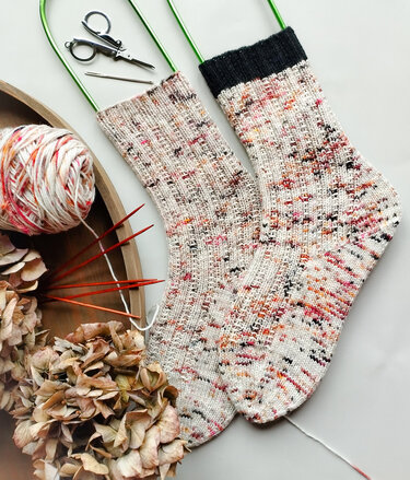 Zwei gestrickte Sprenkelsocken in Creme mit dunklem Bündchen, Garnknäuel und Nadelspiel