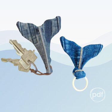 Two denim whale-tail keychains on a pale blue background; one attached to metal keys with a short leather loop, the other secured to a plastic ring with a knotted finish.