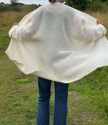 Rückenansicht einer Person in einer cremefarbenen, offen gehäkelten Jacke über blauer Jeans auf einem Weg in grüner Umgebung.