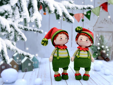 Two crocheted Christmas elf amigurumi dolls in green overalls and red hats on a snowy wooden background