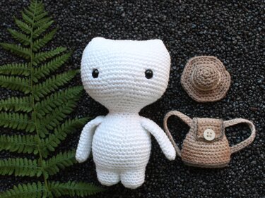 Weißes gehäkeltes Amigurumi-Kätzchen mit schwarzen Augen, brauner Mini-Hut und kleiner Tasche, Farnblatt links