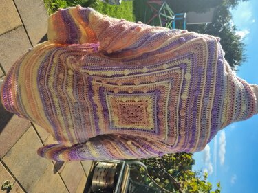 Gehäkelter Seelenwärmer in Pastellfarben mit zentralem Quadrat- und Noppenmuster, im Freien drapiert.