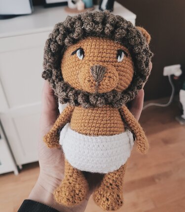 Gehäkelter Löwen-Amigurumi mit brauner Mähne, orangefarbenem Körper und weißer Windel, gehalten in einer Hand.