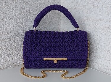 Rechteckige lila gehäkelte Handtasche mit Klappe, kurzem Henkel und goldfarbener Kette vor strukturierter Wand