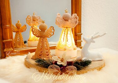 Drei gehäkelte Engel in Gold- und Cremefarben, einer beleuchtet, auf einer dekorativen Holzscheibe neben einer weißen Rehfigur
