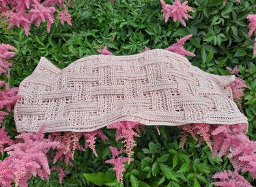 Pale blush crocheted triangle shawl with basketweave textured stitch laid over pink flowering plants