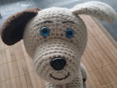 Nahaufnahme eines gehäkelten Hundekopfes in hellbeige mit einem braunen Ohr, blauen Augen und schwarzer Nase auf Holzuntergrund.