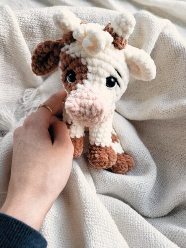 Nahaufnahme einer gehäkelten braun-weißen Kuh-Amigurumi mit schwarzen Augen und rosa Schnauze auf cremefarbener Decke.