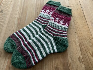 Handgestrickte Socken in Grün, Rot und Weiß mit Häuser- und Streifenmuster auf Holz.