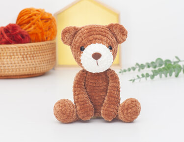 Brown crocheted amigurumi teddy bear sitting on a white surface with a basket of yarn and a yellow prop behind it