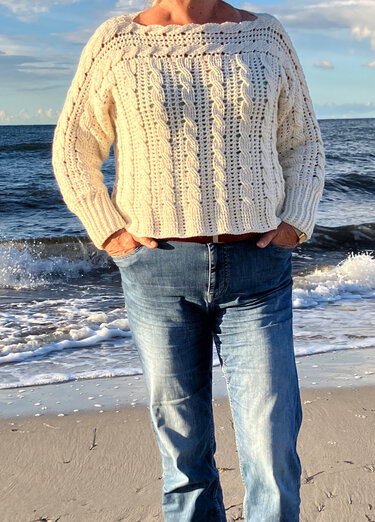Person am Strand trägt cremefarbenen, locker gehäkelten Pullover mit Zopfmuster und Jeans