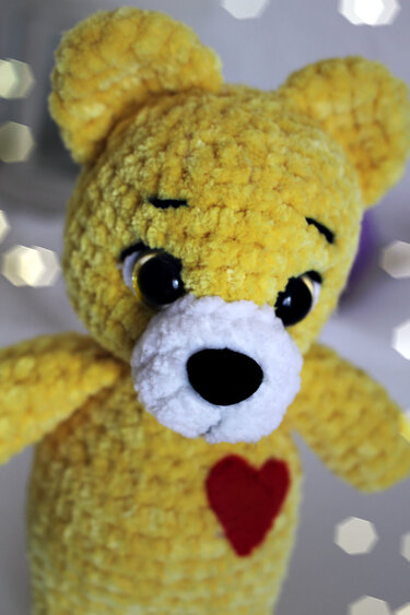 Close-up of a yellow crocheted amigurumi teddy bear with white snout, large black eyes, black nose, and a red heart on its chest