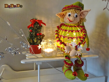 Gehäkelte Elf-Figur mit langen Beinen und Glöckchen neben einem Kerzenglas und Mini-Weihnachtsbaum