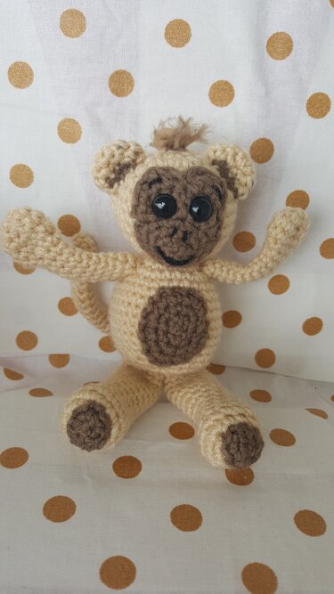 Gehäkelter Amigurumi-Affe in Beige und Braun, sitzend vor weißem Hintergrund mit goldenen Punkten