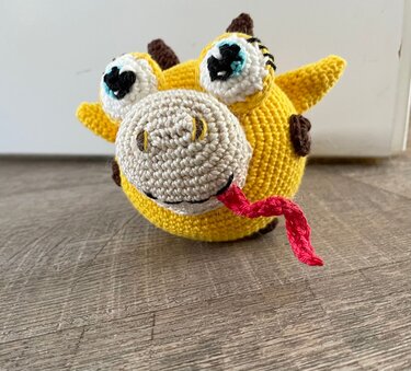 Rundes, gelbes Häkel-Amigurumi mit großen Augen und rosa Zunge auf Holzboden; als Deko oder beschwerter Türstopper nutzbar.