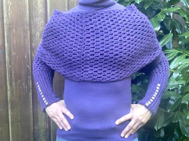 Person trägt ein gehäkeltes lila Schultercape mit offenem Muster über einem gerippten Langarmshirt mit Knopfdetails an den Ärmeln.