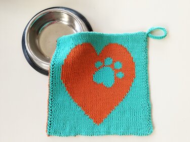 Turquoise knitted potholder with an orange heart and paw motif, corner hanging loop, metal bowl at top left