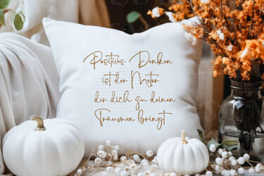 Nahaufnahme eines weißen Kissens mit dem Zitat ‚Positives Denken ist der Motor der dich zu deinen Träumen bringt‘, umgeben von weißen Kürbissen und herbstlicher Deko.