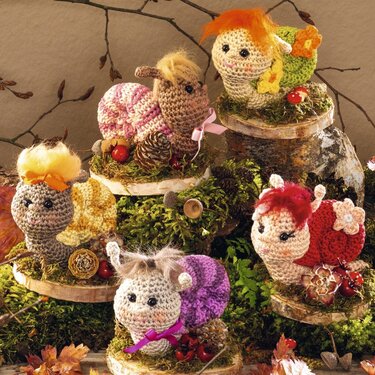 Fünf gehäkelte Amigurumi‑Schnecken mit bunten Schalen, Garnhaar und Moos auf Holzscheiben