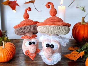 Zwei gehäkelte Pilz-Gnome mit orangefarbenen Hüten und Bärten, umgeben von Kürbissen und Herbstblättern.
