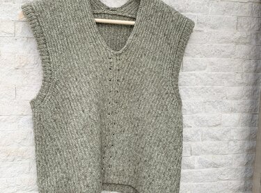 Gestrickte ärmellose Damenweste in Grüngrau-Melange mit V-Ausschnitt, aufgehängt an einem Holzbügel vor heller Steinwand.