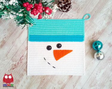 Quadratischer gehäkelter Topflappen mit Schneemanngesicht, türkisfarbener Mütze, oranger Nase und Aufhängeöse