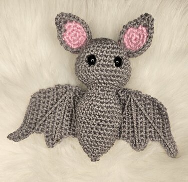 Gehäkelte graue Fledermaus‑Amigurumi mit rosa Ohren und schwarzen Augen auf weißem Kunstfell
