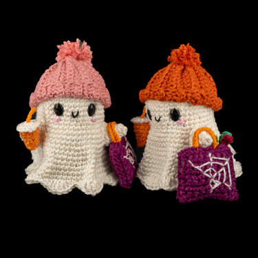 Zwei weiße gehäkelte Geister-Amigurumi mit rosa und orange Mütze, schwarzen Augen, rosa Wangen, orange Süßigkeitseimern und violetten Taschen