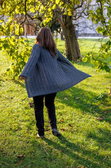 Back view of a woman wearing a long gray ribbed crochet jacket walking on grass beneath a tree