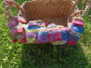 Sechs bunte, gestrickte Fäustlinge für Kinder und Babys in einem geflochtenen Korb auf Rasen.