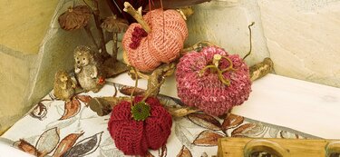 Drei gehäkelte Kürbisse in Rot- und Rosatönen mit Aststielen und kleinen Deko-Igeln auf gemustertem Tischläufer