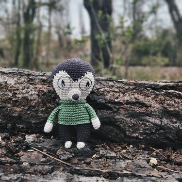 Gehäkeltes Igel-Amigurumi mit runder Brille und grünem Pullover auf einem Baumstamm vor Waldhintergrund.