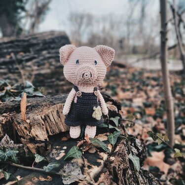 Rosa gehäkeltes Amigurumi‑Schwein mit Latzhose auf moosigem Baumstumpf im Wald