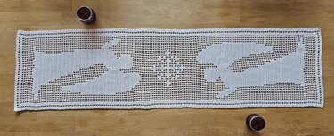 Weißer, handgehäkelter Tischläufer in Filethäkeltechnik mit Rentier- und Sternmotiven auf Holztisch, daneben zwei Kerzengläser.