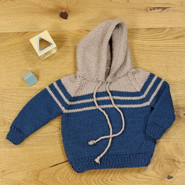 Gestrickter Kinderpullover mit Kapuze, blau unterer Teil, beige Passe mit Streifen, liegend auf Holz