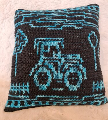 Gehäkeltes quadratisches Kissen mit blauem Traktor‑Motiv und stilisierten Bäumen auf dunklem Grund.