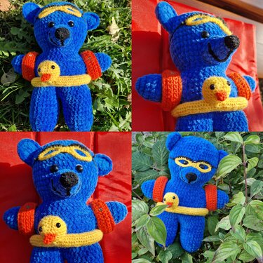 Gehäkelter blauer Teddybär mit gelber Schwimmbrille, orangefarbenen Schwimmflügeln und Schwimmring mit kleiner gelber Badeente.
