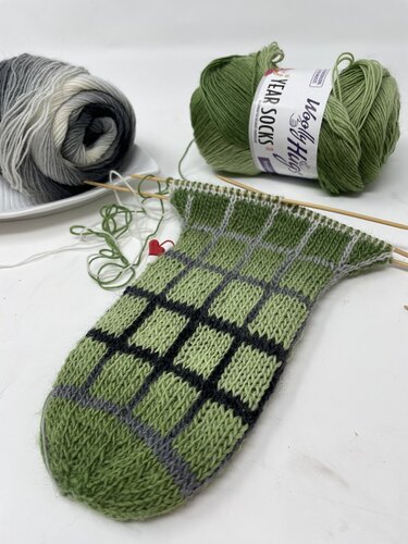 Halbfertige grüne Socke mit schwarz-grauem Karomuster und Garnknäuel
