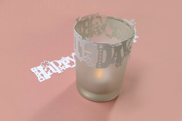 Mattiertes Teelichtglas mit weißer Papiermanschette „Happy Birthday“ und innen leuchtender Kerze