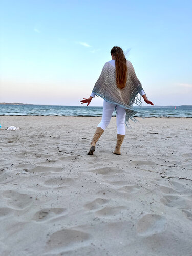 Rückenansicht einer Person am Strand in hellgrauem gehäkelten Poncho mit Fransen